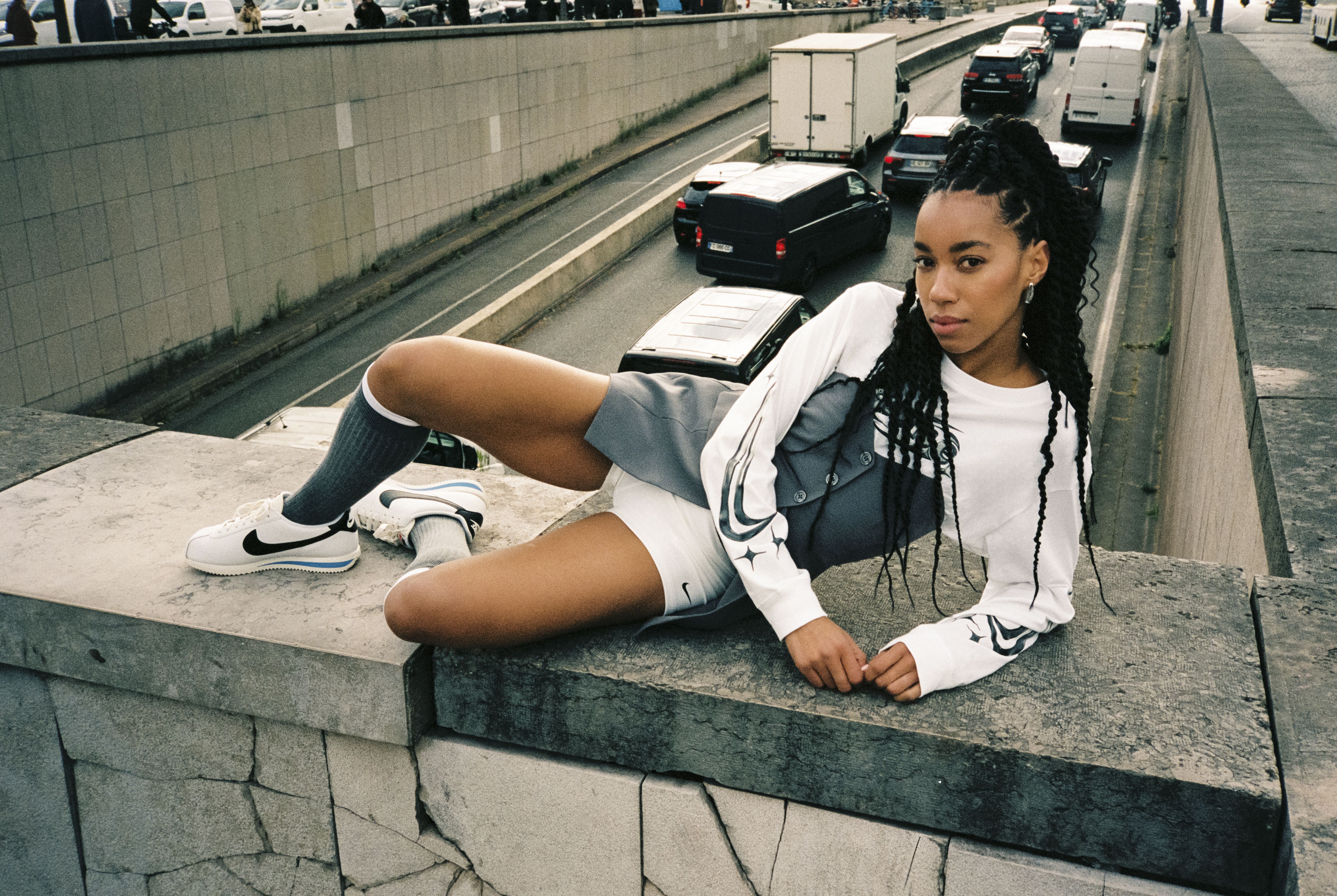 Femeie în bluză albă de la NIKE și cu încălțăminte albă de la NIKE pozează pe un zid de beton, deasupra unei străzi aglomerate